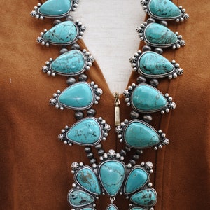 May include: A turquoise squash blossom necklace with silver accents. The necklace features large, teardrop-shaped turquoise stones set in ornate silver settings, strung on a beaded chain. The necklace is displayed against a brown background.