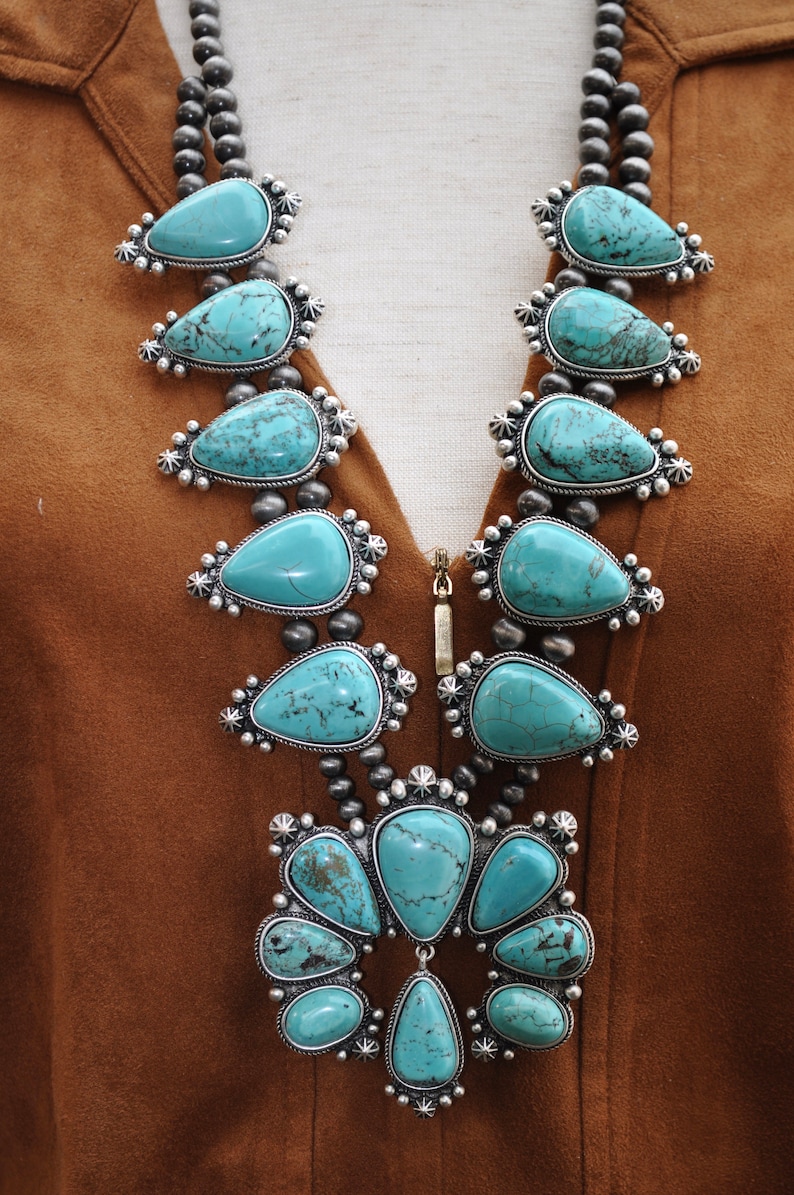 May include: A turquoise squash blossom necklace with silver accents. The necklace features large, teardrop-shaped turquoise stones set in ornate silver settings, strung on a beaded chain. The necklace is displayed against a brown background.