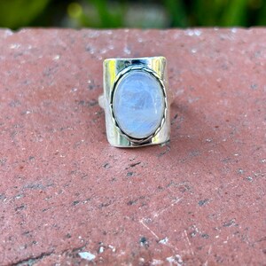 Puede incluir: Un anillo de plata con una gran piedra de luna ovalada engastada en un bisel rectangular. El anillo tiene una banda texturizada.