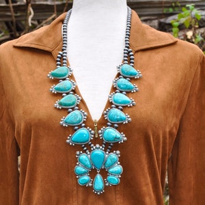 May include: A turquoise and silver statement necklace with a bib design. The necklace features large, teardrop-shaped turquoise stones set in silver, strung on a beaded chain. The necklace is displayed on a brown garment.