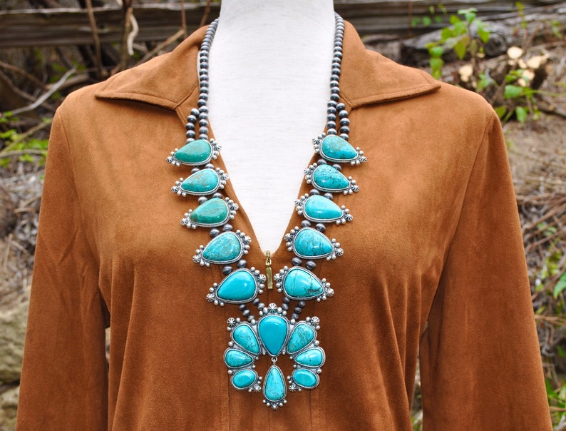 May include: A turquoise and silver statement necklace with a bib design. The necklace features large, teardrop-shaped turquoise stones set in silver, strung on a beaded chain. The necklace is displayed on a brown garment.