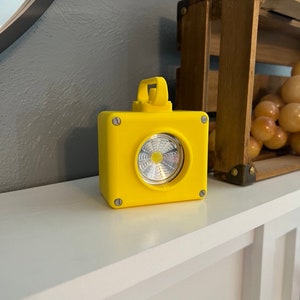 May include: Bright yellow, cube-shaped LED light with a handle. The light has a circular, silver-colored lens and is secured with four screws. The light is sitting on a white surface.