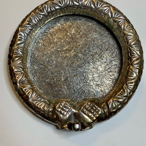 May include: A silver circular tray with an intricate embossed design. The tray has a raised rim and a decorative handle in the shape of two hands clasped together.