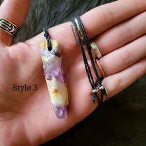 Peut inclure: Pendentif en cristal d'am&eacute;thyste brut sur un collier en cordon noir. Le cristal pr&eacute;sente des nuances de violet, de blanc et de jaune. Le collier est pr&eacute;sent&eacute; sur une main, avec des bagues aux doigts. Le texte "Style 3" est visible.