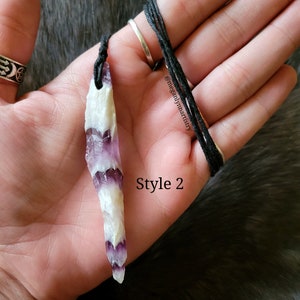 Peut inclure: Un collier pendentif en cristal naturel avec un cordon noir. Le cristal est blanc et violet, avec un motif en couches. Le texte "Style 2" est visible. Le collier est tenu dans une main.