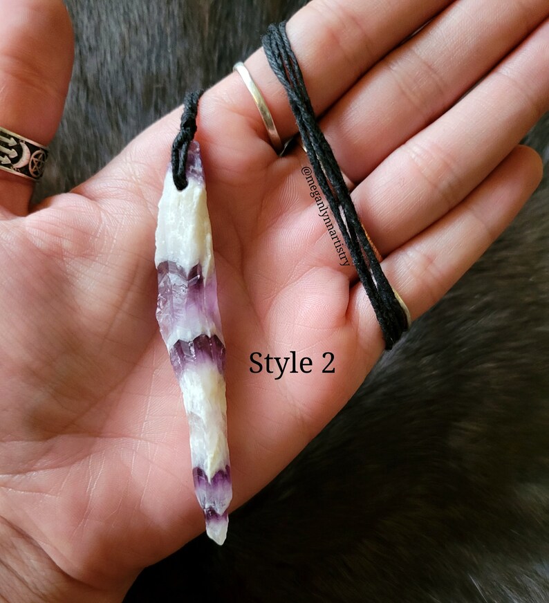 Peut inclure: Un collier pendentif en cristal naturel avec un cordon noir. Le cristal est blanc et violet, avec un motif en couches. Le texte "Style 2" est visible. Le collier est tenu dans une main.