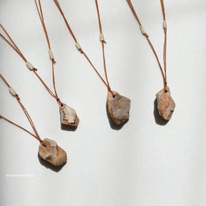 May include: Five stone pendant necklaces with brown cords. Each pendant is a unique, irregularly shaped stone in shades of beige, brown, and gray. The cords are knotted at the top and bottom of each stone. The necklaces are displayed against a white background.