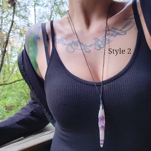 Peut inclure: Un long collier pendentif avec un cristal blanc et violet. Le cristal est suspendu &agrave; un cordon noir. Le texte "Style 2" est visible sur l'image. Le collier est port&eacute; sur un d&eacute;bardeur noir c&ocirc;tel&eacute;.