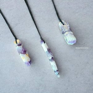 Peut inclure: Trois pendentifs en cristal brut avec des cordons noirs. Les cristaux pr&eacute;sentent un m&eacute;lange de teintes violettes, blanches et jaunes. Chaque pendentif est unique en forme et en taille, mettant en valeur les variations naturelles.