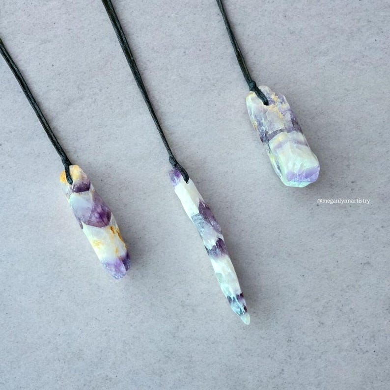 Peut inclure: Trois pendentifs en cristal brut avec des cordons noirs. Les cristaux pr&eacute;sentent un m&eacute;lange de teintes violettes, blanches et jaunes. Chaque pendentif est unique en forme et en taille, mettant en valeur les variations naturelles.
