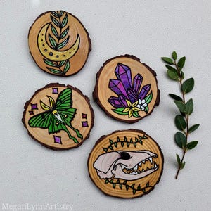 Pode incluir: Quatro bases de madeira pintadas à mão. Os desenhos incluem uma lua crescente e folhas, uma mariposa verde, cristais roxos e uma caveira. As bases são redondas com grão de madeira natural. Cada base tem cerca de 8 cm de diâmetro.