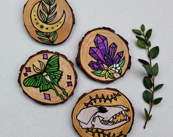 May include: Four hand-painted wooden coasters. Designs include a crescent moon and leaves, a green moth, purple crystals, and a skull. The coasters are circular with a natural wood grain. Each coaster is approximately 8 cm in diameter.