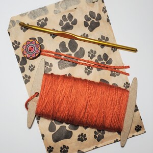 May include: A brown paper bag with a paw print pattern. The bag contains a skein of orange yarn, a wooden yarn needle, and a crochet hook. There is a colorful button with a floral design on the bag.