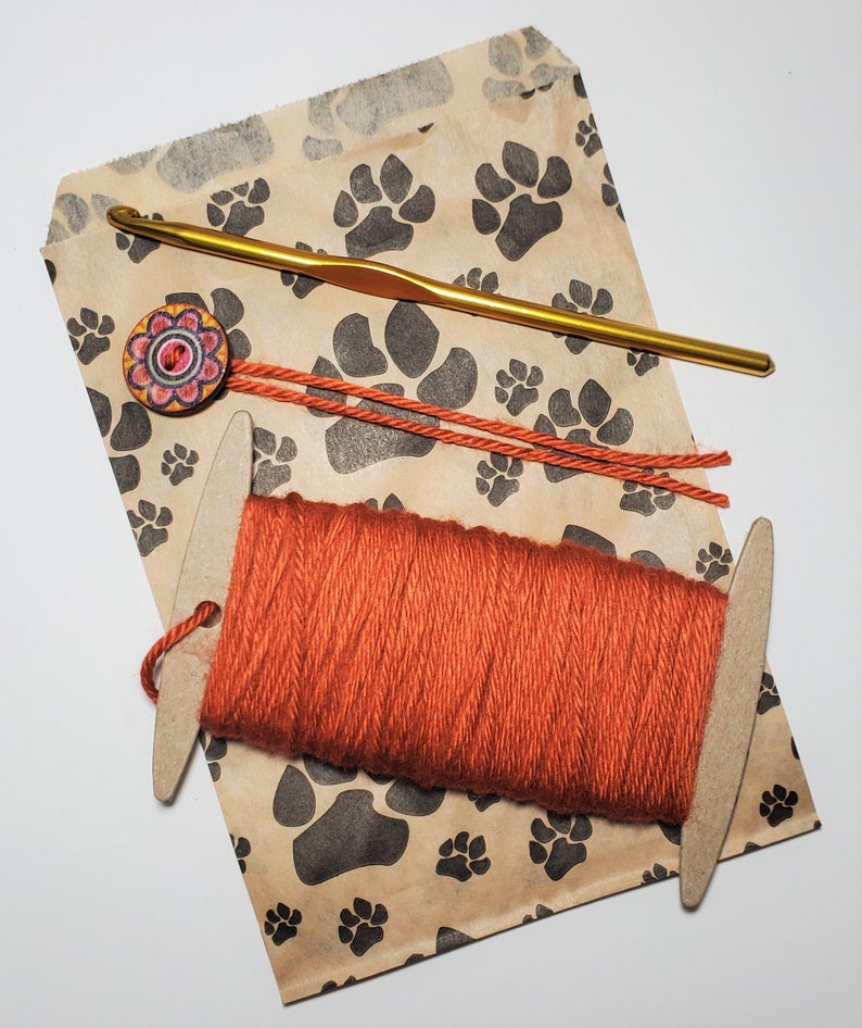 May include: A brown paper bag with a paw print pattern. The bag contains a skein of orange yarn, a wooden yarn needle, and a crochet hook. There is a colorful button with a floral design on the bag.