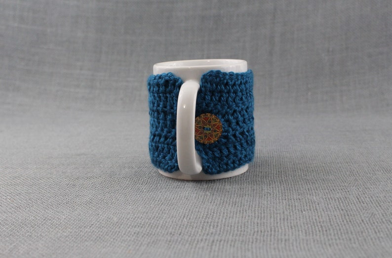 May include: A white ceramic mug with a teal blue crocheted cozy. The cozy has a decorative button with a multi-colored design.