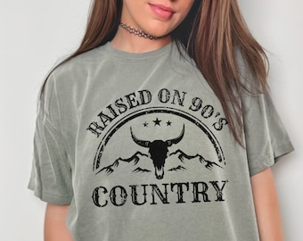 May include: A moss green t-shirt with a black graphic print that says "Raised on 90's Country" with a skull and crossbones and mountains in the design. The brand is Comfort Colors.