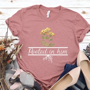 May include: A dusty rose t-shirt with a white graphic of a yellow flower with roots and the text "Rooted in him".