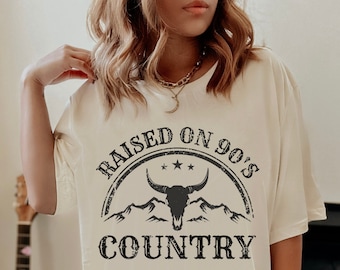 May include: An oversized white t-shirt with a black graphic print that says "RAISED ON 90'S COUNTRY" with a skull and mountain design.