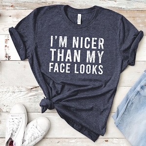May include: Navy blue t-shirt with the white text "I'M NICER THAN MY FACE LOOKS." The shirt is knotted at the waist. White sneakers and blue jeans are visible in the image. Small potted plants are also present.
