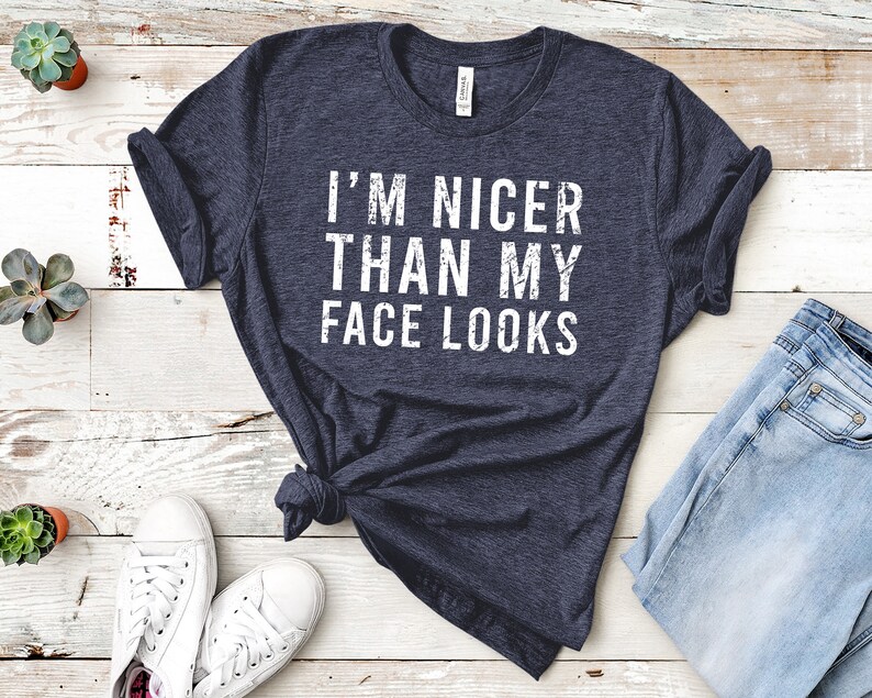 May include: Navy blue t-shirt with the white text "I'M NICER THAN MY FACE LOOKS." The shirt is knotted at the waist. White sneakers and blue jeans are visible in the image. Small potted plants are also present.