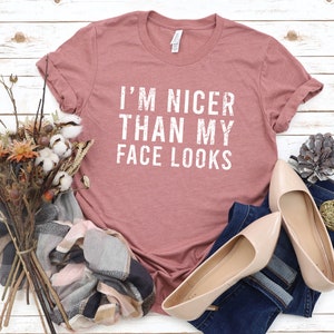 May include: A dusty rose t-shirt with the text "I'M NICER THAN MY FACE LOOKS" in white. The shirt is laid on a white wooden surface, next to a pair of beige flats, a denim pant, and a bouquet of dried flowers.