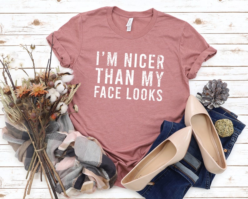 May include: A dusty rose t-shirt with the text "I'M NICER THAN MY FACE LOOKS" in white. The shirt is laid on a white wooden surface, next to a pair of beige flats, a denim pant, and a bouquet of dried flowers.