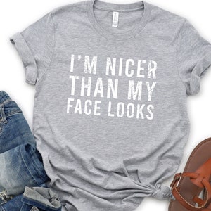 May include: A heather gray t-shirt with the text "I'M NICER THAN MY FACE LOOKS" in white. The shirt is styled with blue jeans and brown sandals. The t-shirt is a casual wear item.