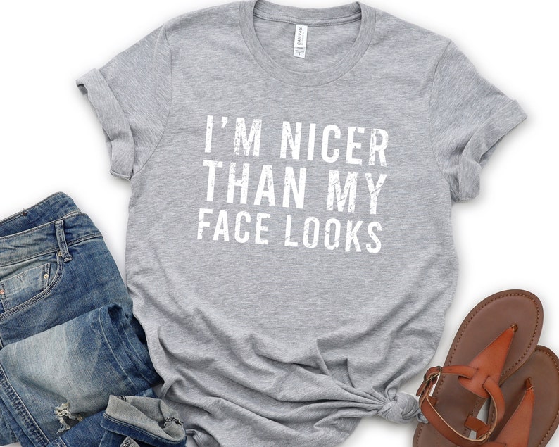 May include: A heather gray t-shirt with the text "I'M NICER THAN MY FACE LOOKS" in white. The shirt is styled with blue jeans and brown sandals. The t-shirt is a casual wear item.