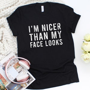 May include: Black t-shirt with the white text "I'M NICER THAN MY FACE LOOKS". The shirt is displayed on a white surface with a magazine and headphones in the foreground. White flowers are in the background.