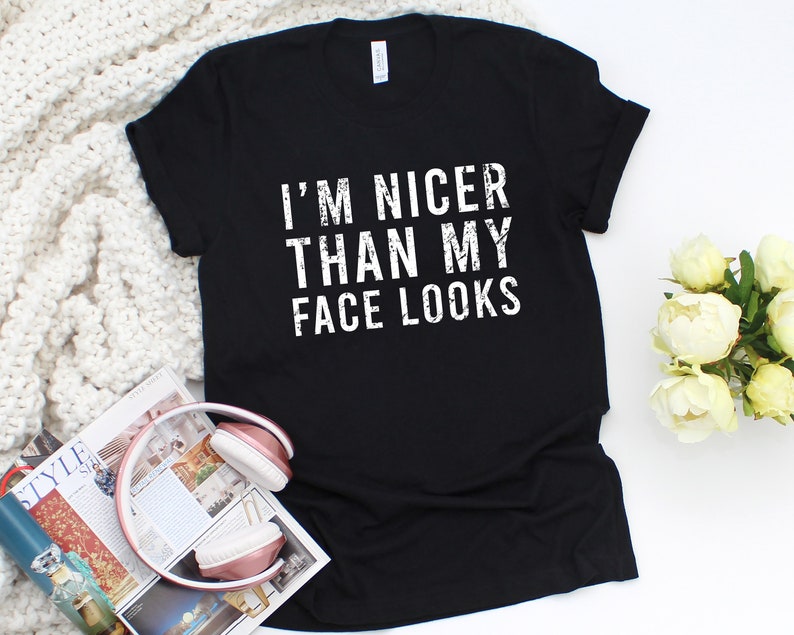 May include: Black t-shirt with the white text "I'M NICER THAN MY FACE LOOKS". The shirt is displayed on a white surface with a magazine and headphones in the foreground. White flowers are in the background.