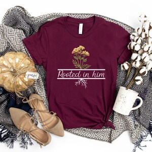 May include: A maroon t-shirt with a white graphic of a yellow flower with roots and the text "Rooted in him".