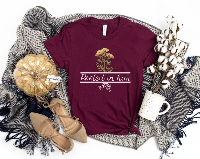May include: A maroon t-shirt with a white graphic of a yellow flower with roots and the text "Rooted in him".