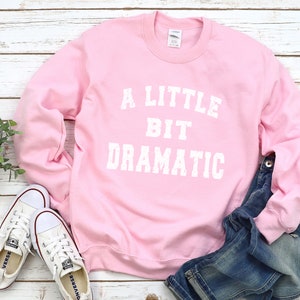 May include: A pink sweatshirt with the text "A Little Bit Dramatic" in white lettering.
