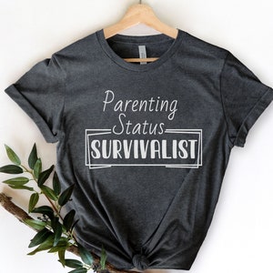K&ouml;nnte beinhalten: Ein dunkelgraues T-Shirt mit dem Text "Parenting Status SURVIVLIST" in Wei&szlig;.