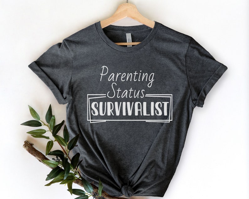 K&ouml;nnte beinhalten: Ein dunkelgraues T-Shirt mit dem Text "Parenting Status SURVIVLIST" in Wei&szlig;.