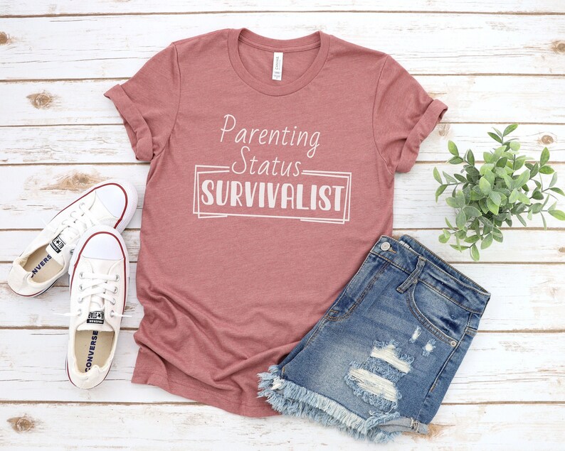 K&ouml;nnte beinhalten: Ein heidefarbenes T-Shirt mit dem Text "Parenting Status Survivalist" in wei&szlig;en Buchstaben.