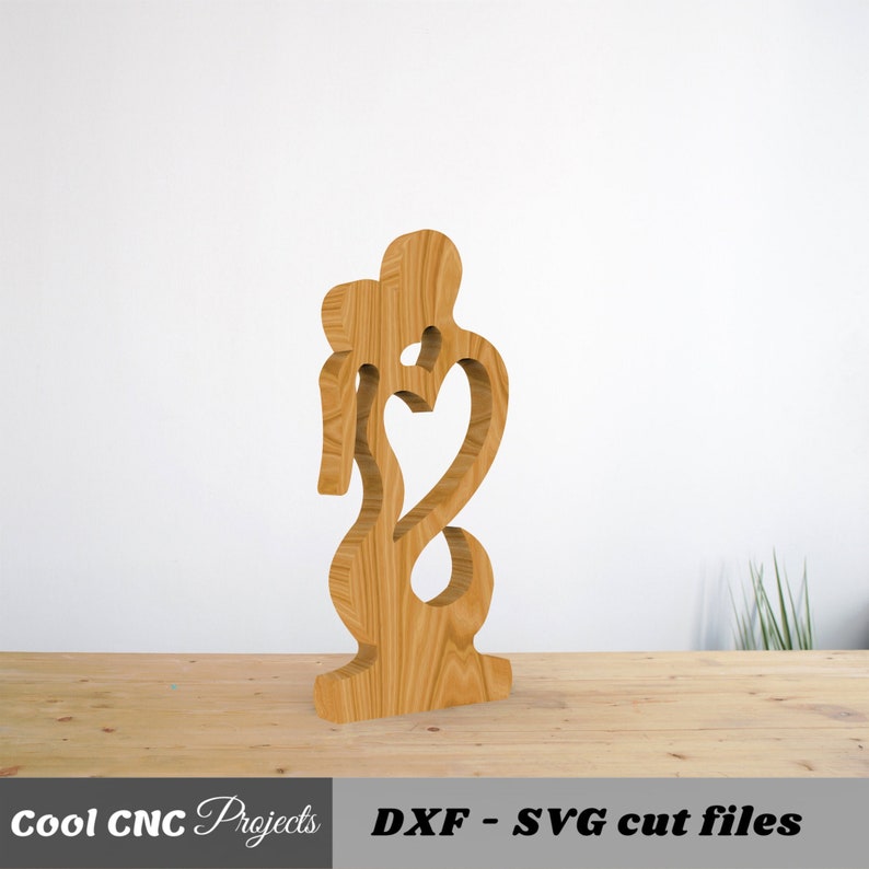 May include: Wooden cutout of two figures forming a heart shape. The figures are standing close together, with one figure's arm around the other's waist. The cutout is a light brown color and has a smooth finish.