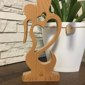 May include: A wooden sculpture of two figures embracing, with a heart cutout in the center. The sculpture is painted in a natural wood color and has the names "Ваня & Радо" engraved on the base.