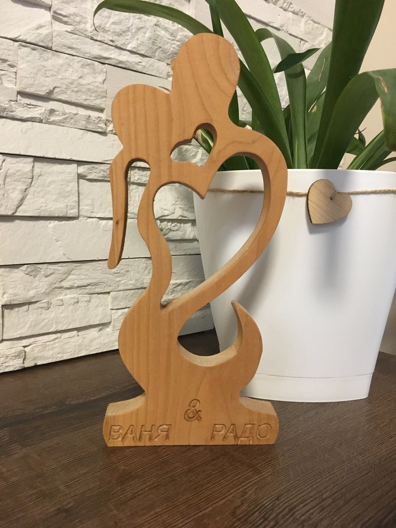 May include: A wooden sculpture of two figures embracing, with a heart cutout in the center. The sculpture is painted in a natural wood color and has the names "Ваня & Радо" engraved on the base.