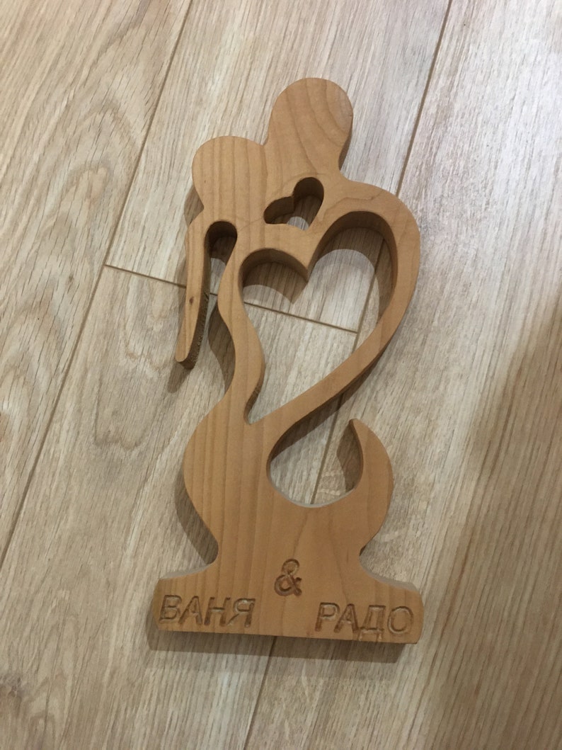 May include: A wooden cutout of two figures embracing, forming a heart shape. The base is engraved with the names "Ваня & Радо".