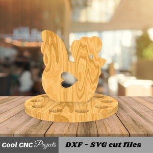 K&ouml;nnte beinhalten: H&uuml;hner-Eierhalter aus Holz mit 12 L&ouml;chern f&uuml;r Eier. Das Huhn steht auf einem runden Sockel mit einem ausgeschnittenen Herzen. Das Holz ist hellbraun.
