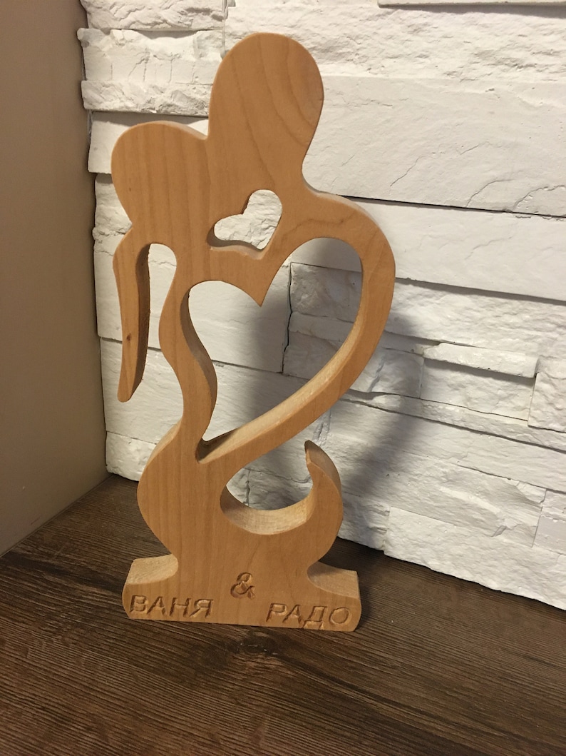 May include: A wooden sculpture of two figures embracing, with a heart cutout in the center. The sculpture is inscribed with the names "Ваня & Радо".