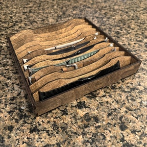 May include: A wooden watch band organizer with multiple watch bands. The organizer is made of layered wood with a dark stain. The watch bands are various colors and materials, including leather and fabric.