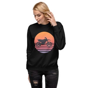 Puede incluir: Sudadera negra con un gráfico de una moto de motocross silueteada contra una puesta de sol. El diseño del atardecer incluye rayas horizontales naranjas, rosas y moradas dentro de un círculo. La sudadera la lleva una persona.