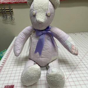 May include: A handmade patchwork teddy bear with a purple quilted body, white fleece ears and feet, and a purple satin bow. The bear has button eyes.
