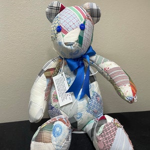 May include: A patchwork teddy bear with blue eyes and a blue ribbon around its neck. The bear is made from a variety of fabrics, including plaid, floral, and solid colors. The bear is sitting on a black surface.