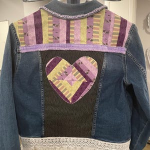 May include: A blue denim jacket with a patchwork heart on the back. The heart is made of purple, yellow, and pink fabric. The jacket has a white lace trim and fringe.
