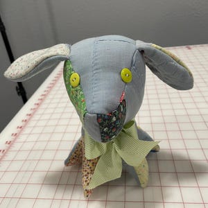 May include: A patchwork fabric dog toy with a light blue body, green and floral patterned ears, and a green and white striped bow. The toy has yellow button eyes.