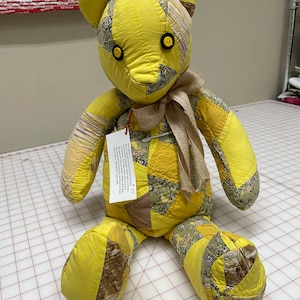 May include: A yellow patchwork teddy bear with brown accents and button eyes. The bear is sitting on a gray and white gridded surface. A tag is attached to the bear's left arm with the text "$7.75".
