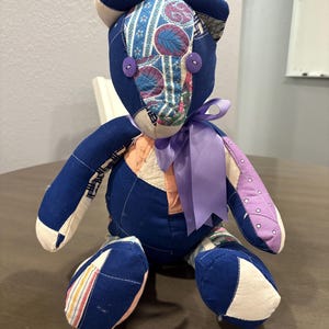 May include: A handmade patchwork teddy bear with a blue body, patterned face, and purple ribbon. The bear features various fabric patterns and is adorned with two purple button eyes. A unique, handcrafted plush toy.
